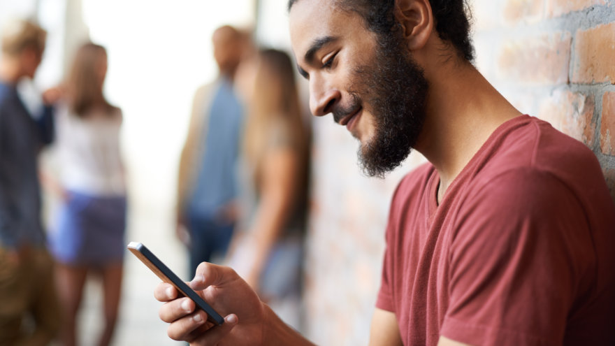 A young man with facial hair softly smiling as he looks down at a message on his phone. A group of his friends can be seen in the background.