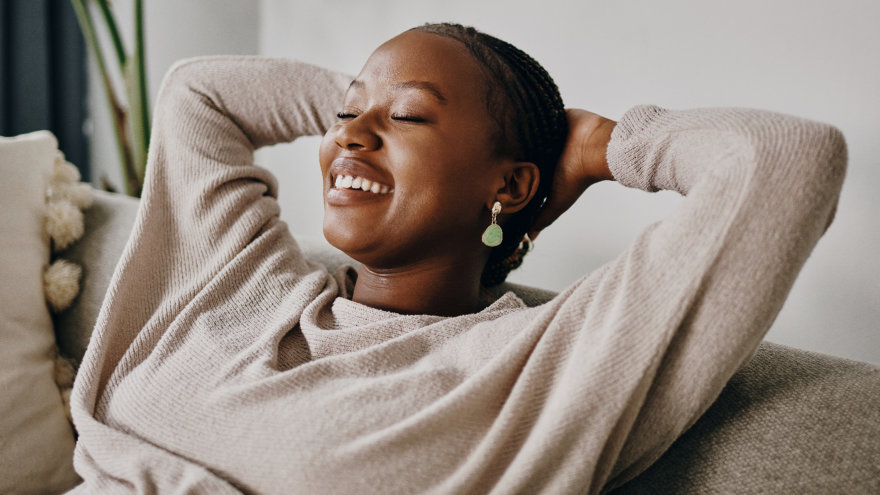 Woman lounging back on her couch with her hands behind her head. Her eyes are closed with a gentle smile on her face, she looks at rest and content.