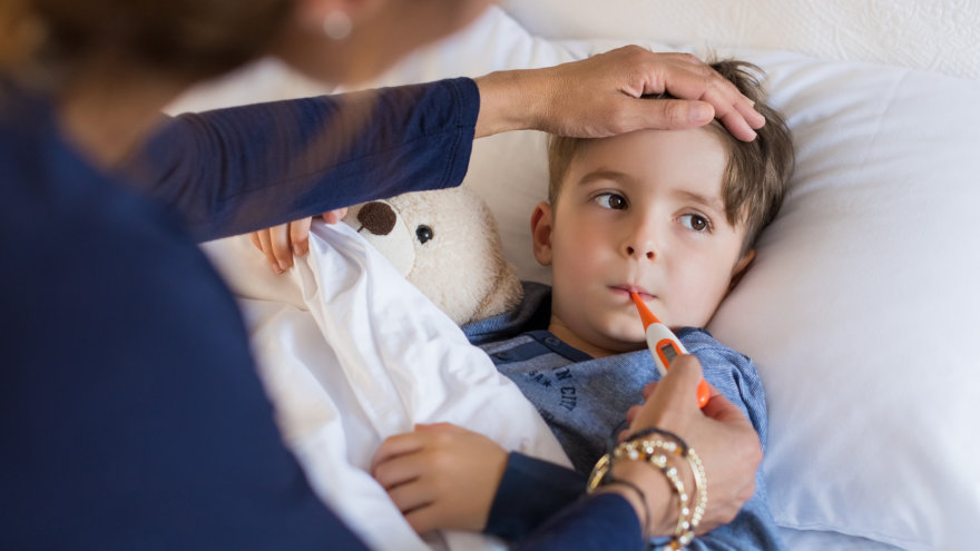 A mother with her hand pressed against her young son's forehead as she takes his temperature. Her son is bundled up under his covers, holding his teddy bear.